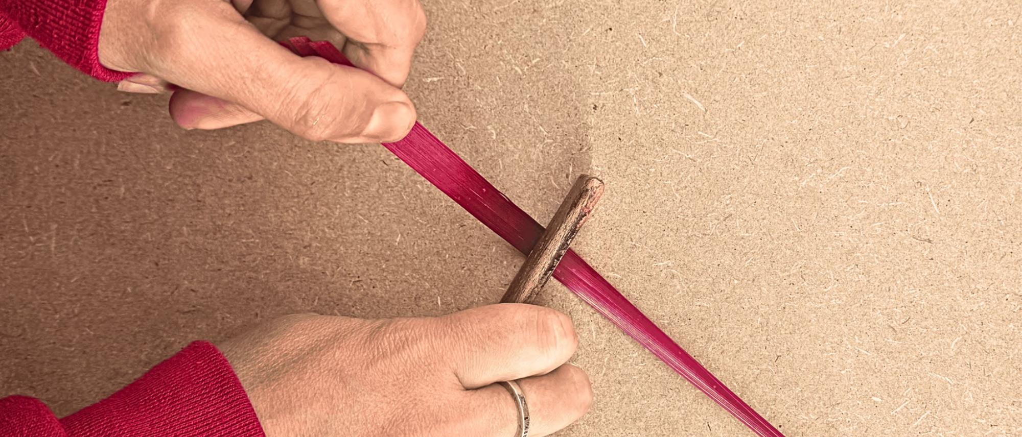 Deux mains fendent un brin de paille de seigle au couteau, sur un plan de travail en bois.